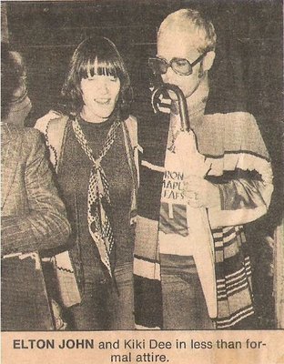 John, Elton (+ Kiki Dee) / Elton Holding Umbrella, Wearing Toronto Maple Leafs T-Shirt | Magazine Photo with Caption (1976)