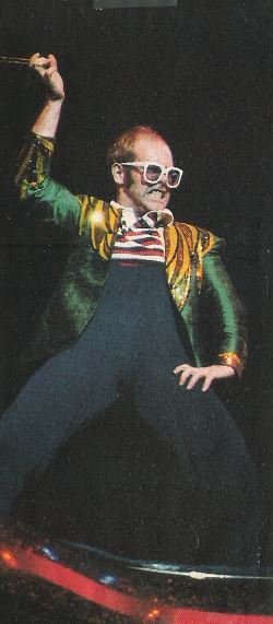 John, Elton / On Top of Piano, Twirling Necklace, Left Hand On Hip, Green-Gold Jacket / 1976