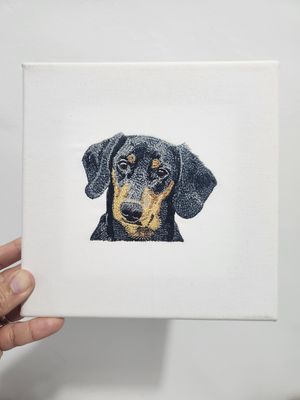 Embroidered black and tan dachshund pet portrait on a small white stretched canvas, held by a hand.
