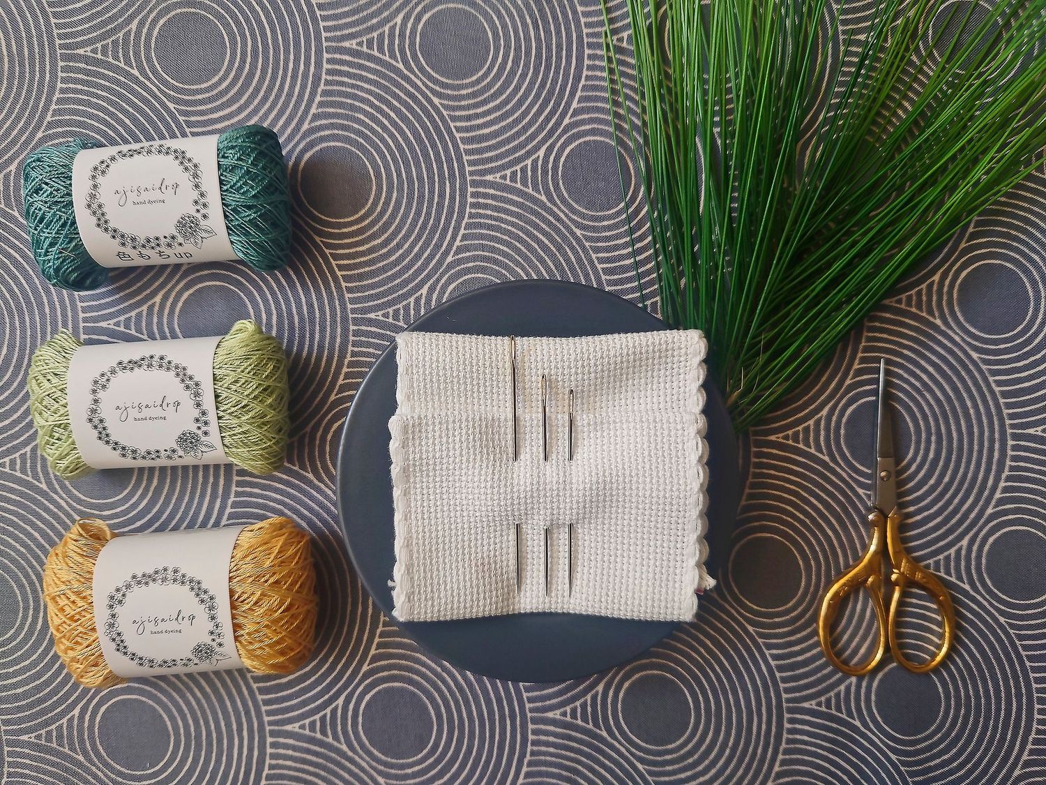 Was ist Sashiko? - Japanische Stickerei links - japanische Sashiko-Stickgarne, im HIntergrund japanischer Stoff, in der MitteSashiko-Sticknadeln, rechts -Stickschere