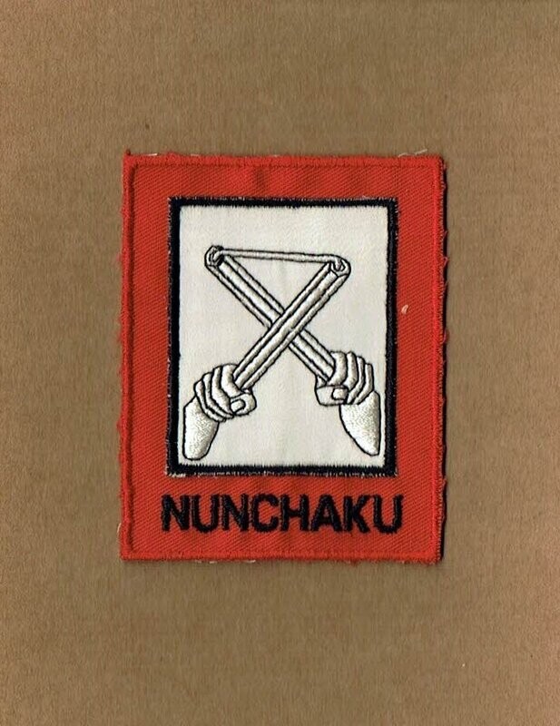 Patch/Écusson Broderie - Représentation de l'Arme Patch/Écusson Broderie - Représentation de l'Arme "Nunchaku" en Blanc dans un Rectangle sous un Deuxième Rectangle sous Fond Rouge Écrit "Munchaku", Dimension 7 x 8.5 cm