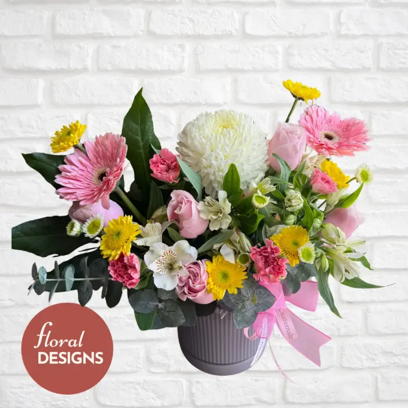 Arreglo floral Siena con rosas rosadas, gerberas, crisantemos y alstroemerias en base decorativa con listón rosa