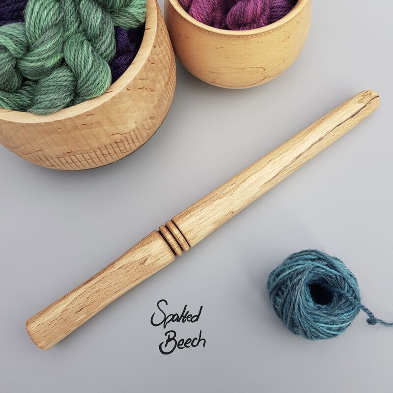 Wooden nostepinne (yarn winder) turned from spalted beech on pale grey surface. To bottom right is a small ball of blue yarn that has been wound with the winder. To the top left are glimpses of 2 wood turned bowls holding mini skeins of yarn.