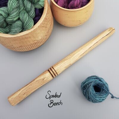 Wooden nostepinne (yarn winder) turned from spalted beech on pale grey surface. To bottom right is a small ball of blue yarn that has been wound with the winder. To the top left are glimpses of 2 wood turned bowls holding mini skeins of yarn.