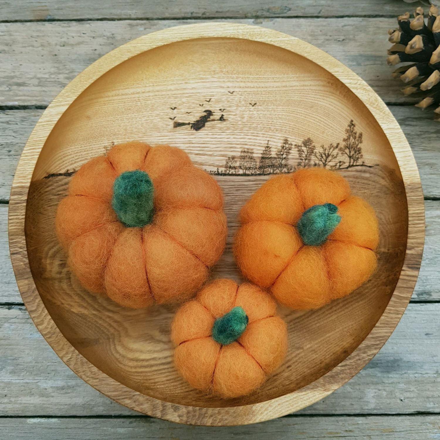 A wooden bowl on a weathered wooden surface, holding three, orange needle felted pumpkins. There is a glimpse of a pine cone to the top right corner.