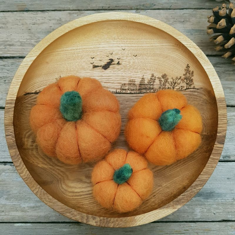 A wooden bowl on a weathered wooden surface, holding three, orange needle felted pumpkins. There is a glimpse of a pine cone to the top right corner.