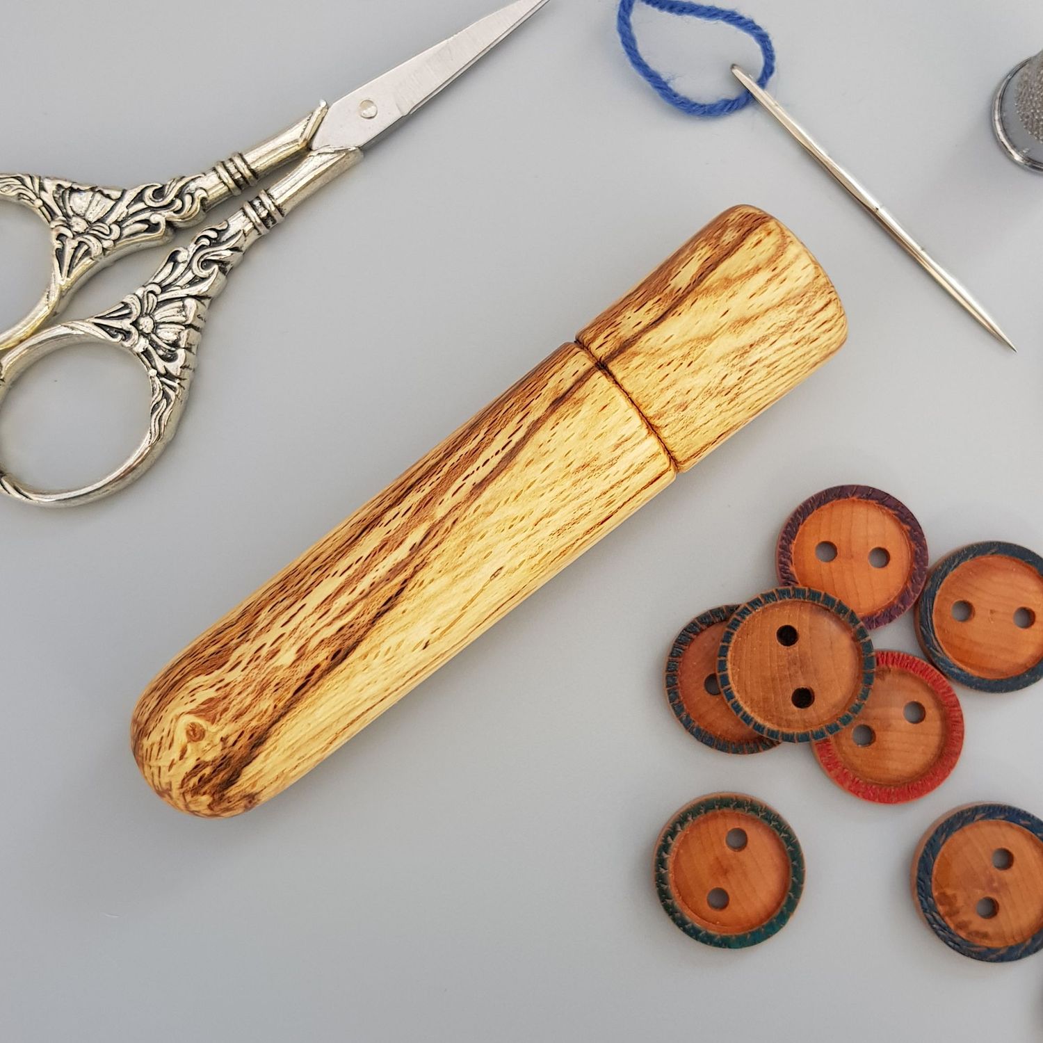 Wooden needle case turned from spalted beech on pale grey background with some wooden buttons, a darning needle holding blue yarn and some embroidery scissors around the case.