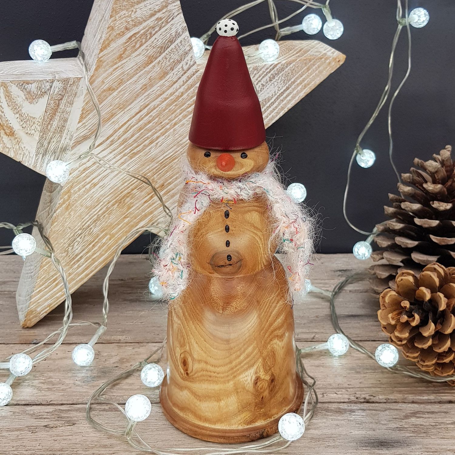 Wooden Snowman 27 Wooden snowman Christmas decoration with a large carrot shaped nose, 24cm tall by 10cm wide (9.5"x 4") turned from elm. Wearing a red Santa shaped hat decorated with white pompom and black dots and a white, with green, orange and red threads, hand-knitted scarf. Behind the snowman is a wooden star, some pine cones and white christmas lights, against a dark grey background.