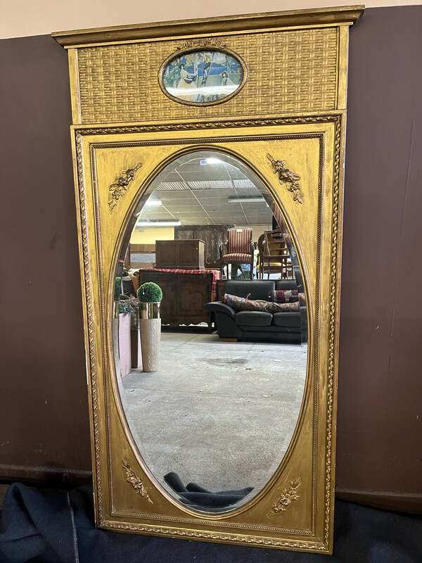 TRUMEAU en bois doré à glace ovale surmontée d'une reproduction représentant trois personnages cueillant des pommes. Dim.: 148 x 81 cm. Quelques manques, accidents et restauration à l'arrière.