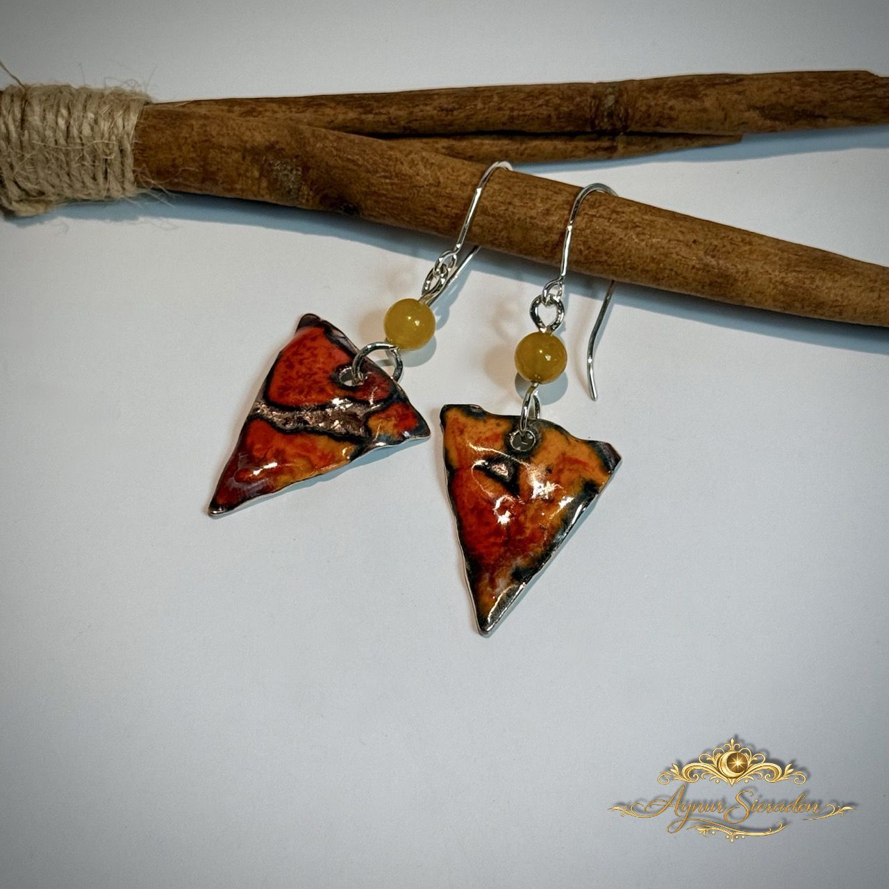 Handgemaakte geëmailleerde oorbellen “Kind van het Vuur” in warme rood- en aardetinten, hangend aan houten tak 