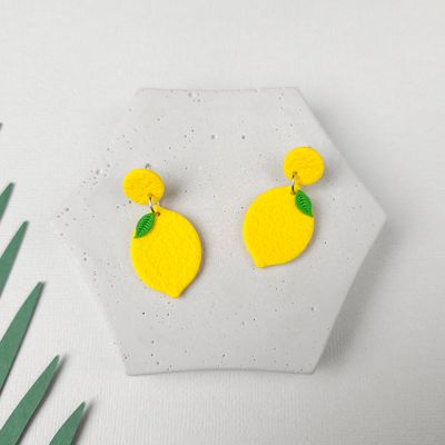 A pair of yellow lemon-shaped earrings with green leaves. The earrings are on a grey hexagonal surface.