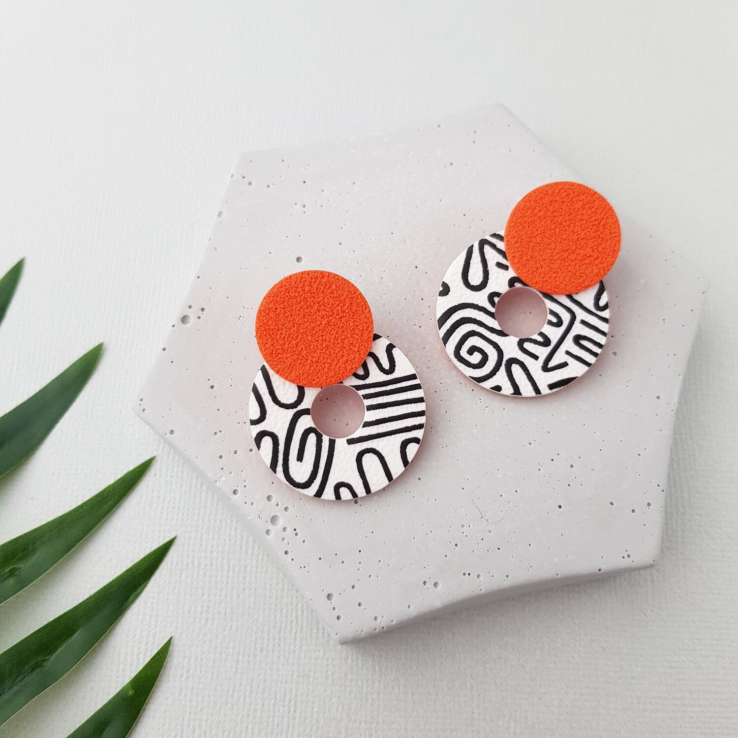A pair of earrings made with a white, circular hoop base with black abstract designs and a textured orange circle on top. 