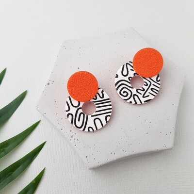 A pair of earrings made with a white, circular hoop base with black abstract designs and a textured orange circle on top. 