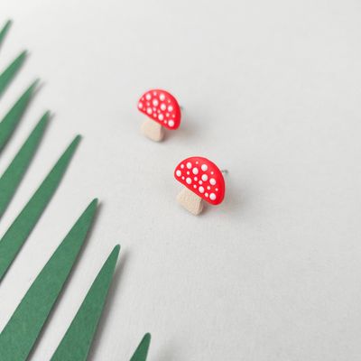 A pair of small mushroom stud earrings. Each earring has a red cap with white spots and a beige stem.