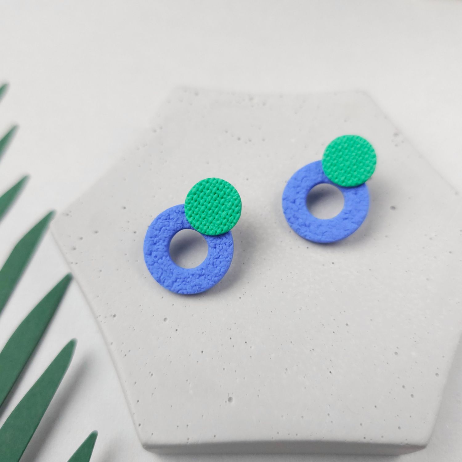 A pair of blue and green stud earrings, which feature a small green circle on top of a larger blue circle hoop at the bottom.
