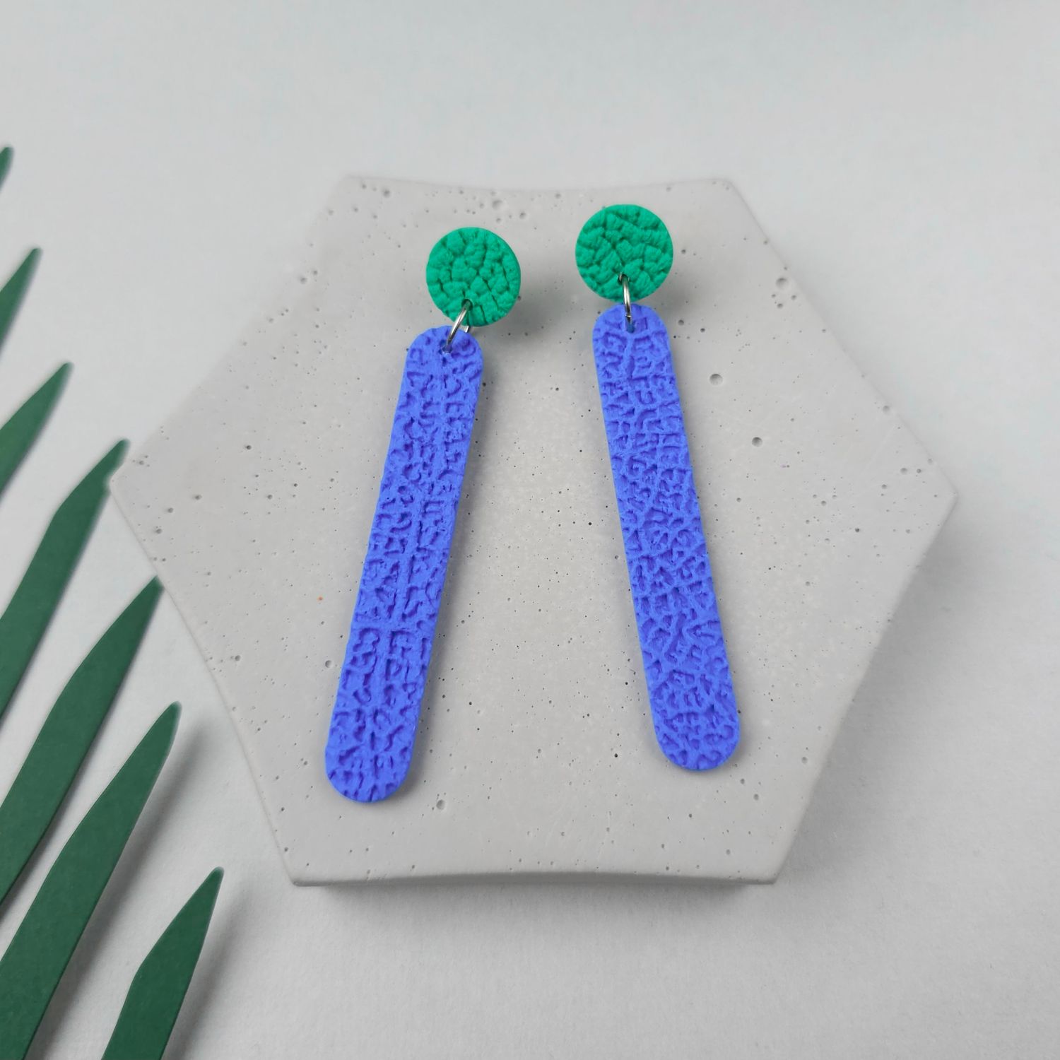 A pair of long earrings with a textured, blue bar and a small, textured, green circle at the top.