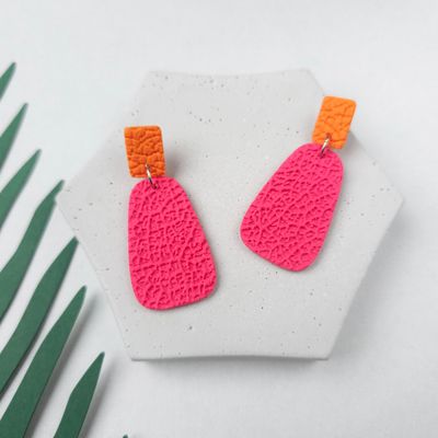 A pair of pink and orange statement earrings with a textured finish. The earrings are on a grey hexagonal surface.