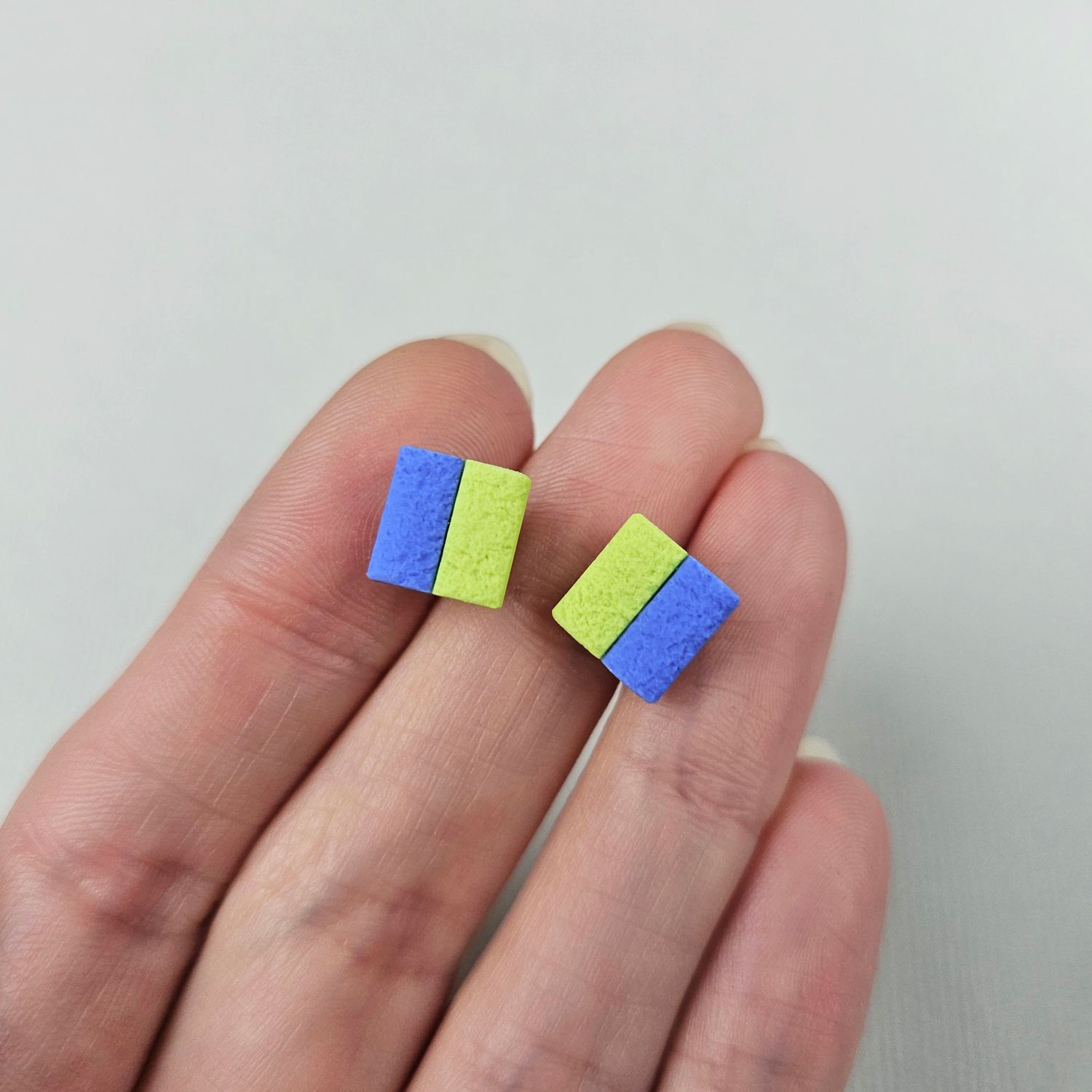 A pair of square earrings with a light green and blue colour scheme, placed on a hand for size reference.