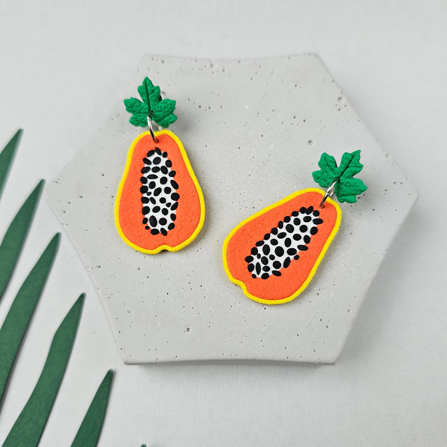 A pair of papaya earrings with green upper leaves, black seeds, and a yellow outline. The earrings are made of polymer clay.