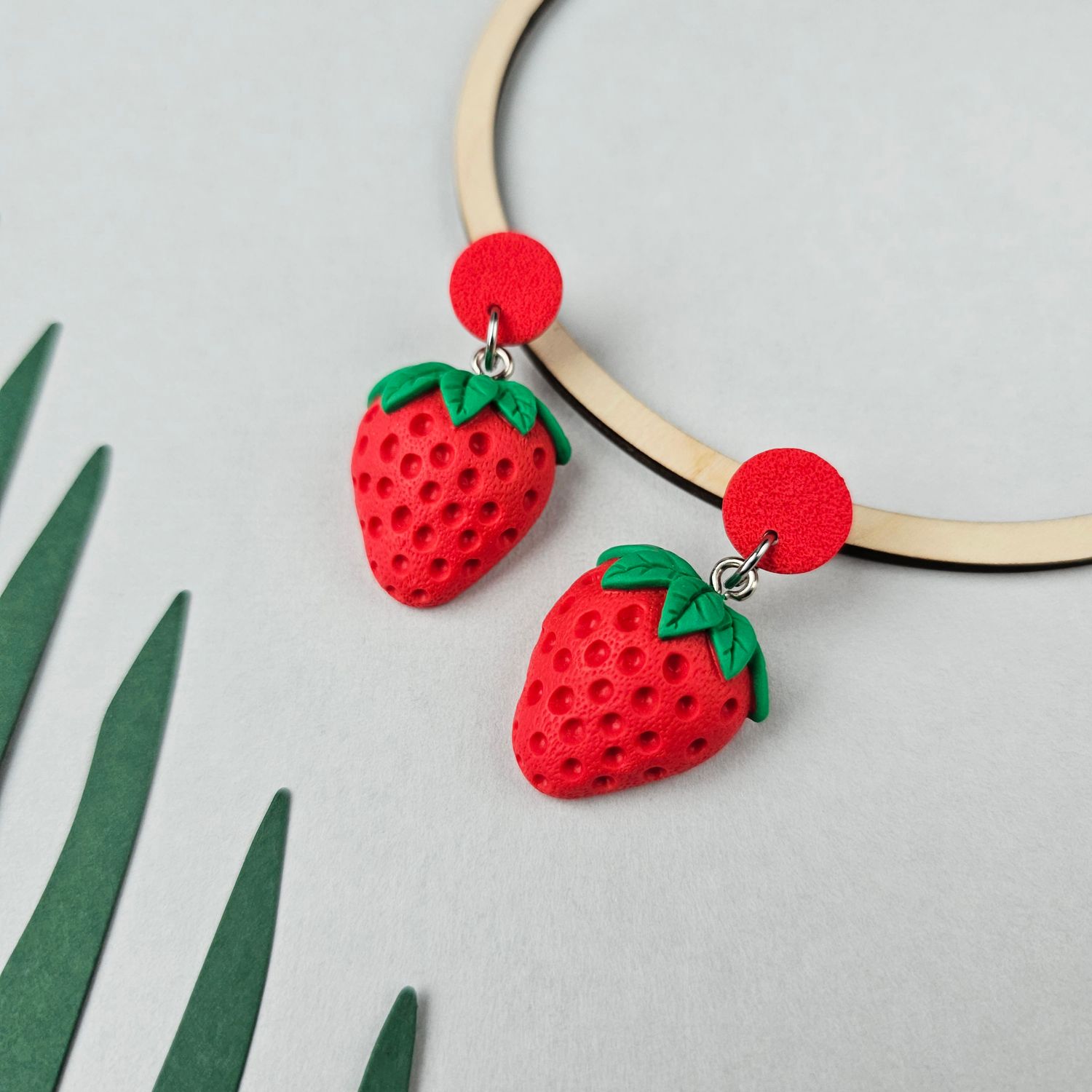A pair of earrings which has a bright red strawberry design with green leaves and a red circular upper stud