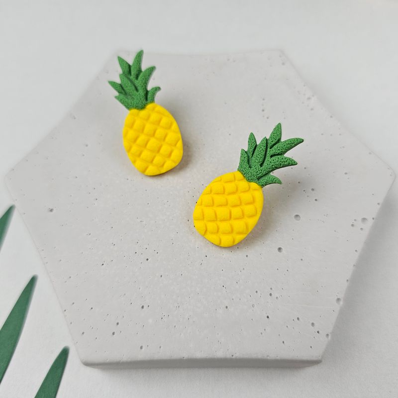 A pair of yellow pineapple earrings with green leaves. The earrings are on a grey textured surface.