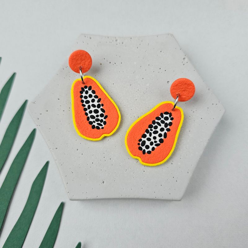 A pair of papaya-shaped earrings with black seeds, orange and white flesh, and yellow outlines. 