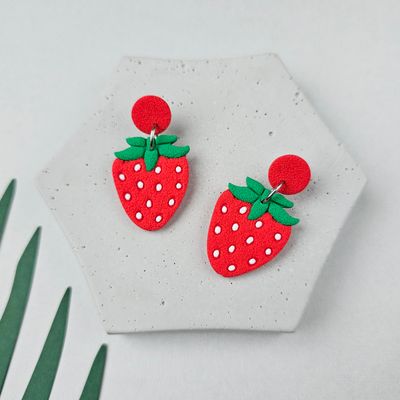 A pair of red and green strawberry earrings with white seeds. The earrings are on a grey hexagonal surface.