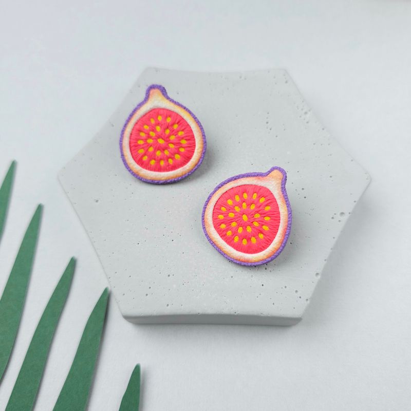 A pair of earrings shaped like a fig, with a pink middle and yellow seeds. The earrings are on a grey hexagonal surface.