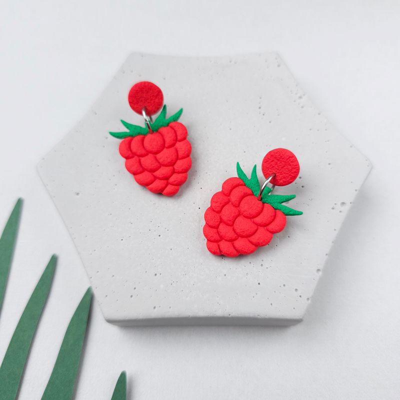 A pair of red raspberry earrings with green leaves and a red upper bead. The earrings are on a grey background.