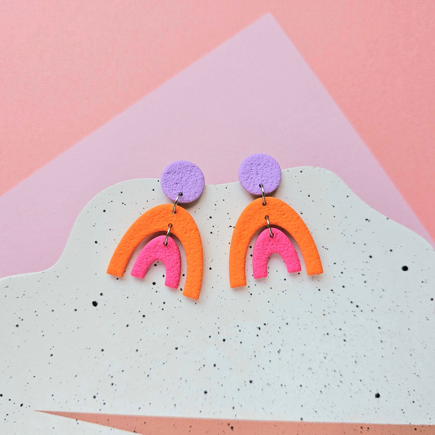 A pair of colourful, abstract earrings that feature a textured, purple circle at the top, and an orange and a pink arch below