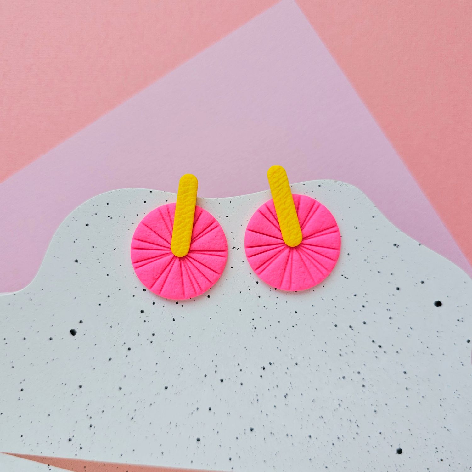 A pair of vibrant pink and yellow geometric earrings that are round with a yellow bar extending from the centre.