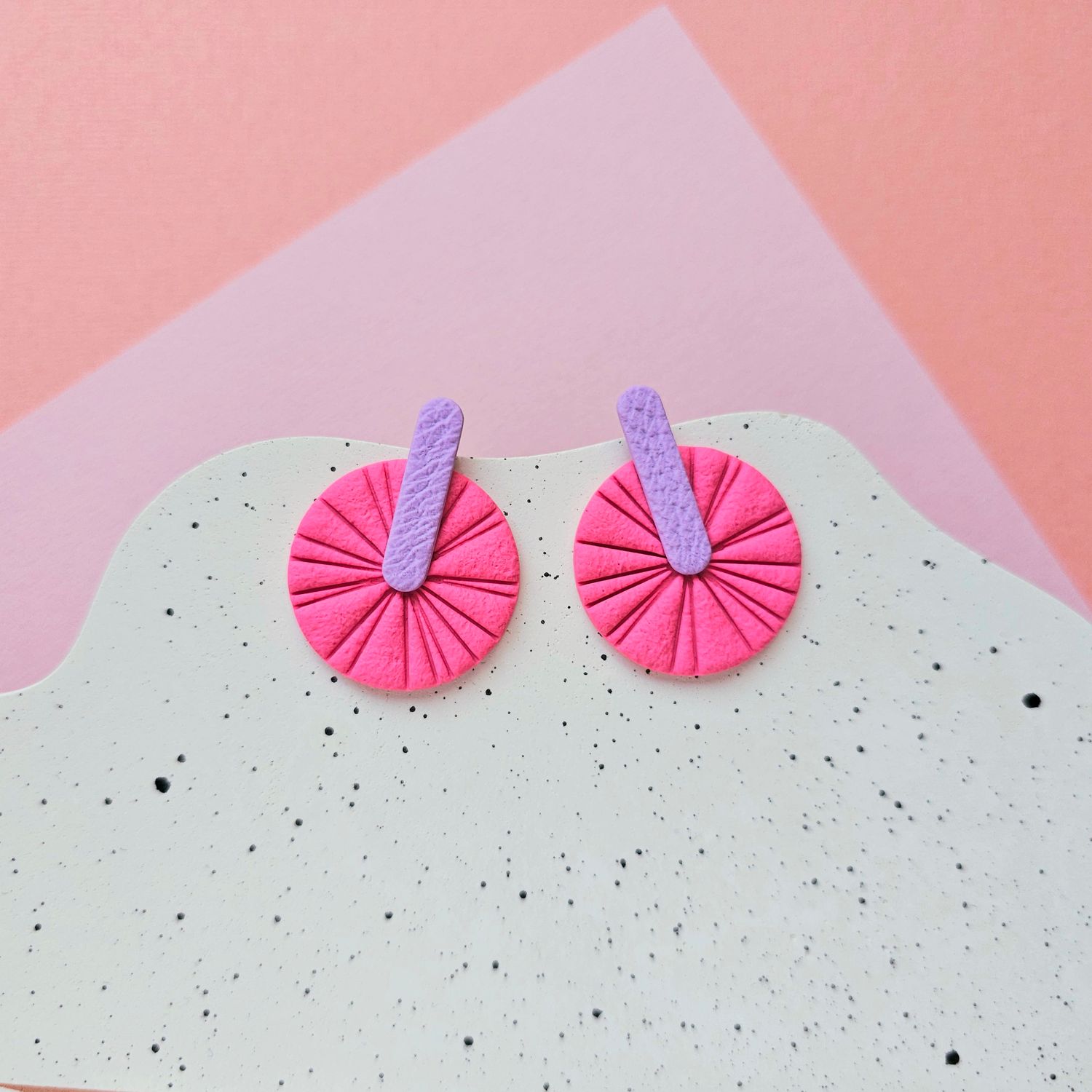 A pair of pink and purple geometric earrings that are round with a textured surface and a purple bar across the centre.