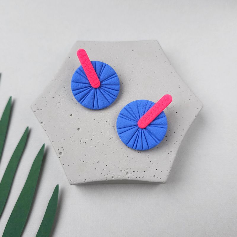 A pair of blue and pink geometric earrings. They are round with a textured surface and a pink bar extending from the centre.