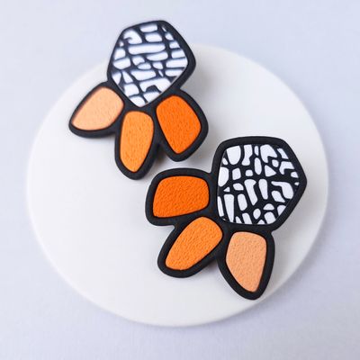 A pair of statement earrings with a geometric design. The earrings are black with white pebble accents and orange petals.
