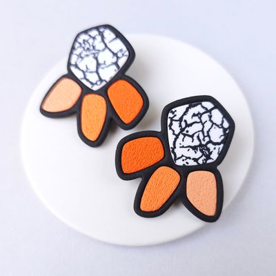 A pair of statement earrings with a geometric design. The earrings are black with white marbled accents and orange petals. 