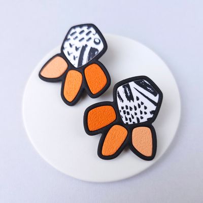 A pair of statement earrings with a geometric design. The earrings are black with white doodle accents and orange petals.