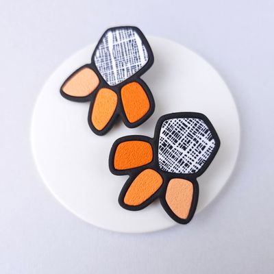 A pair of statement earrings with a geometric design. The earrings are black with white fabric accents and orange petals.