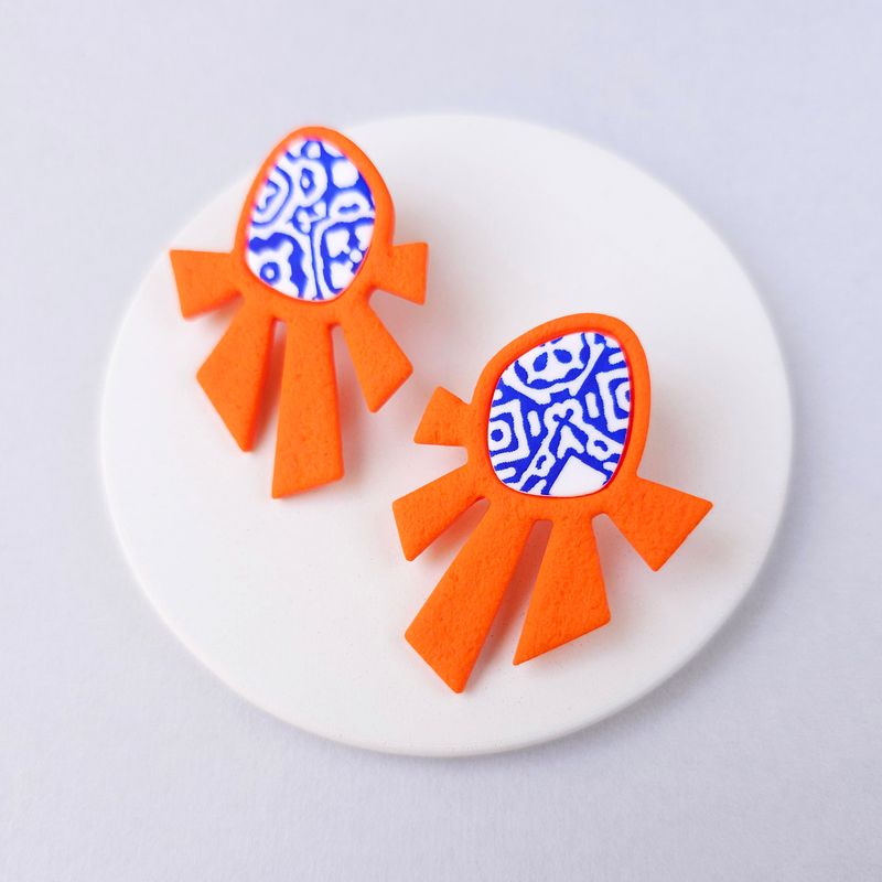 A pair of orange and blue statement earrings that are shaped like a sunburst with a blue and white patterned upper part.