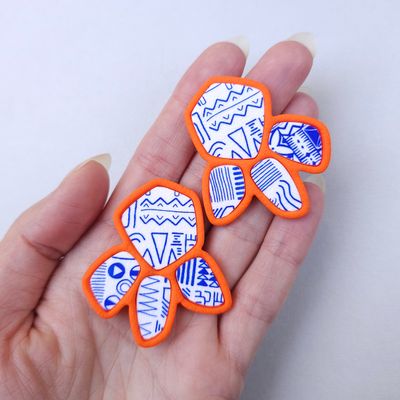 Two abstract, organic-shaped earrings that have a white surface with blue geometric patterns, outlined with an orange border