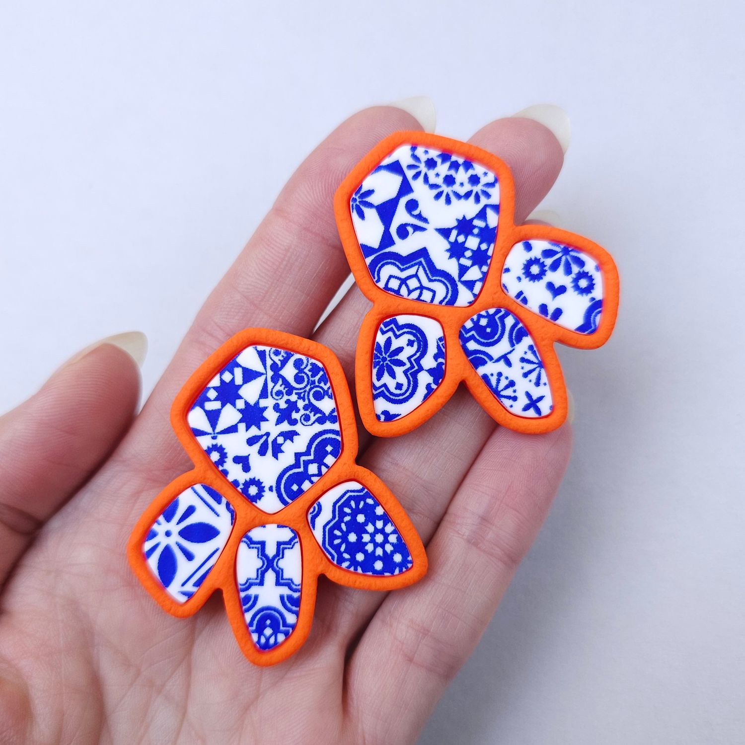 Organic-shaped earrings with a white and blue pattern, edged in orange, are placed on a hand for size reference.