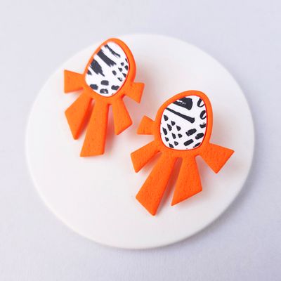 A pair of orange geometric earrings. The earrings are shaped like a sun with a black and white abstract design at the top.