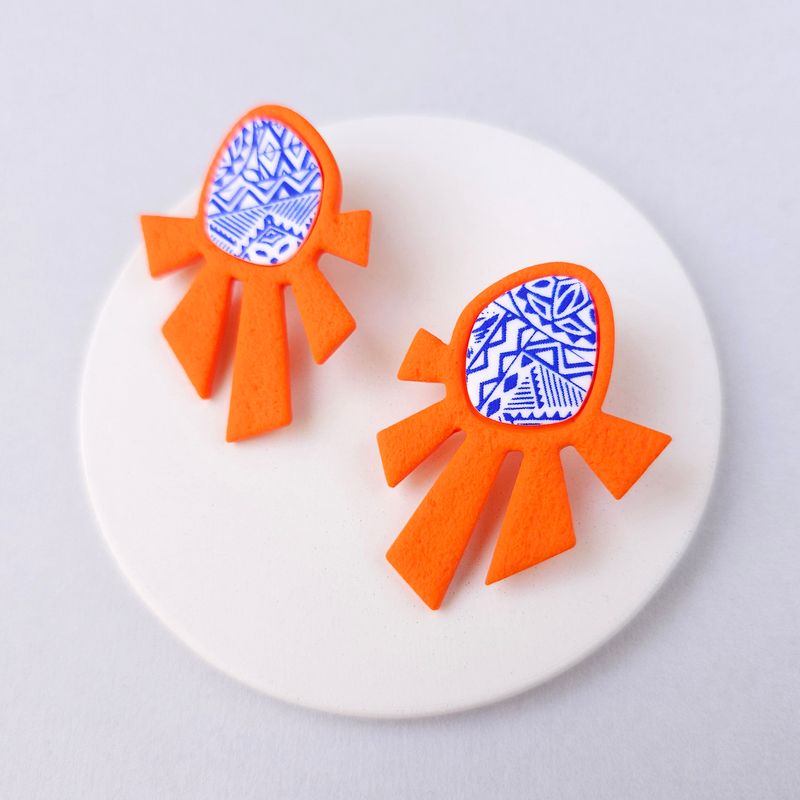 A pair of orange statement earrings with a blue and white geometric pattern at the top.