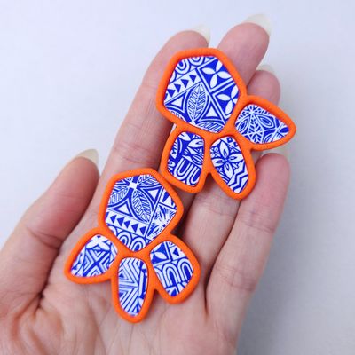 Two organic-shaped earrings with a blue and white tribal pattern. The earrings have an orange border and are worn on a hand
