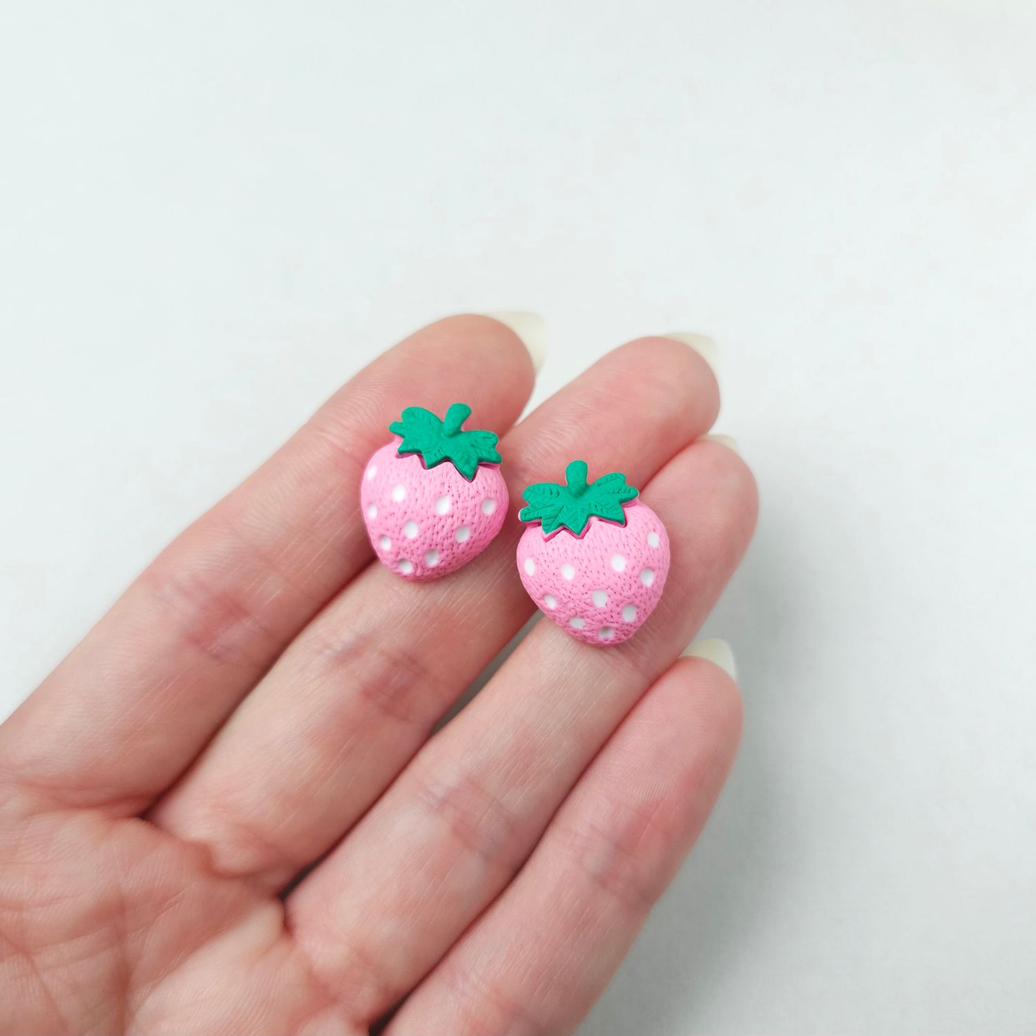 Two small, light pink, strawberry-shaped stud earrings with white seeds and green top leaves.