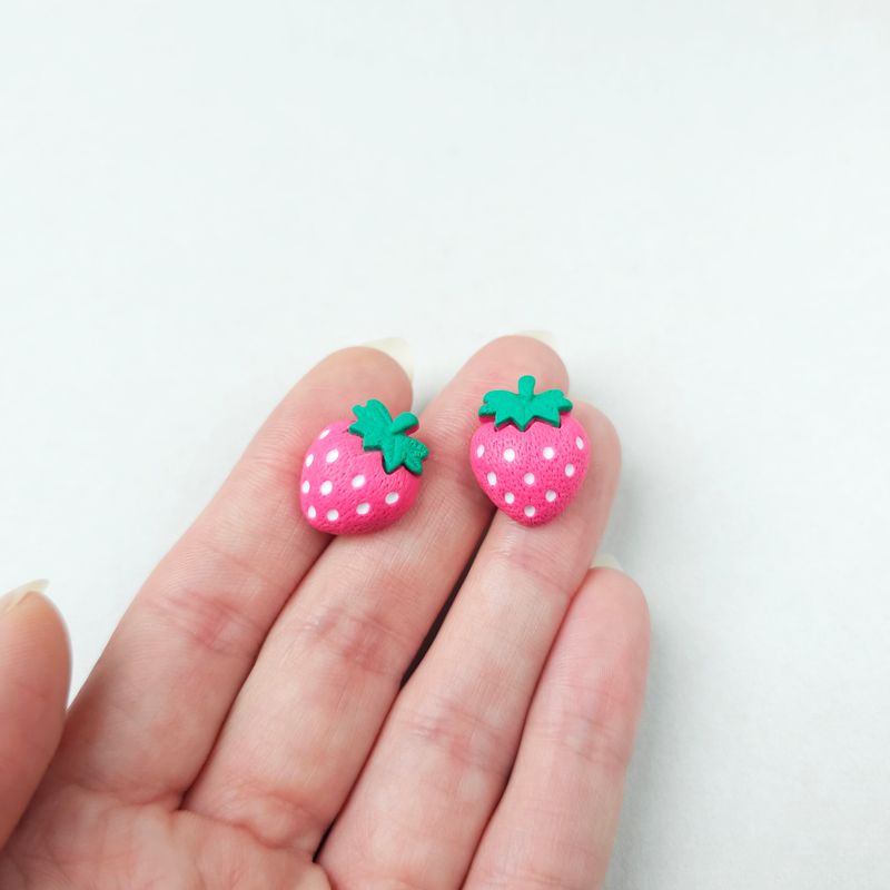Two small, pink strawberry-shaped stud earrings with white seeds and green top leaves. They are positioned on a person's hand