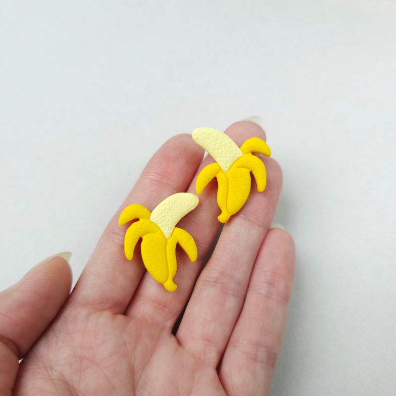 Two yellow banana-shaped studs with a yellow peel. The earrings are set on a hand for size reference.