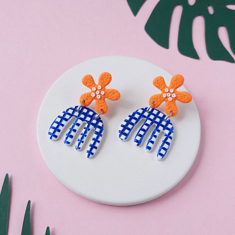 A pair of handmade earrings with orange flower-shaped tops connected to blue and white checkered, curved, organic shapes.