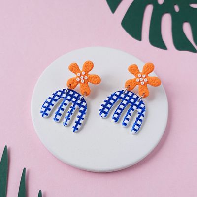 A pair of handmade earrings with orange flower-shaped tops connected to blue and white checkered, curved, organic shapes.