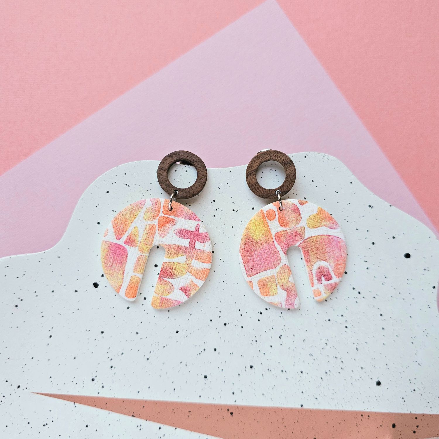 A pair of abstract earrings that have a big white base with a pattern of pink, orange, and yellow and a small top hoop bead.