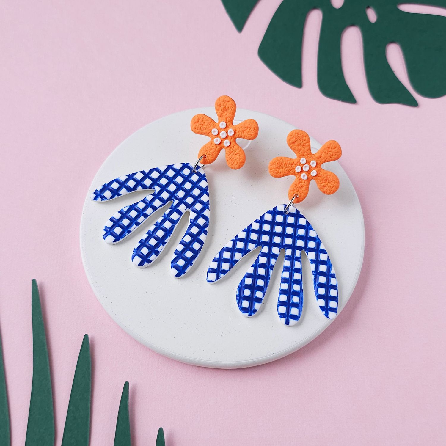 A pair of large earrings with orange flower studs and blue and white checkered leaf-shaped dangles.